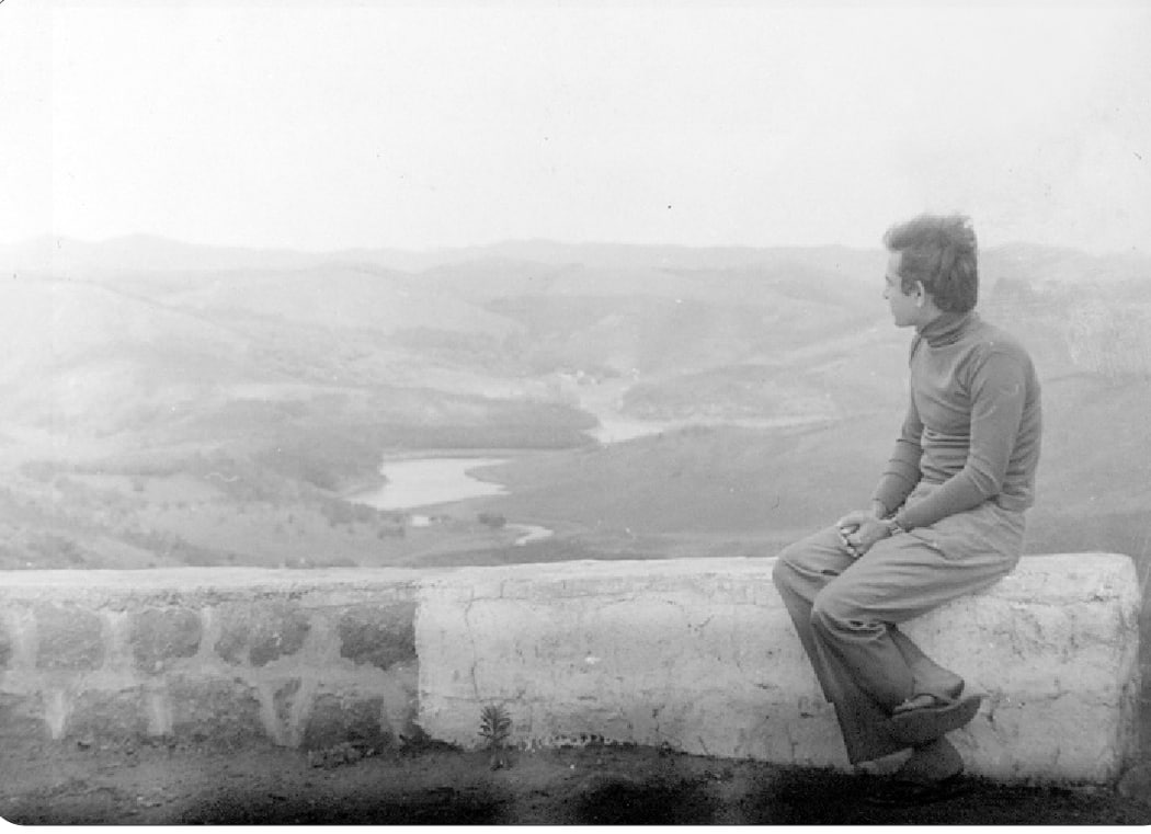 Promesse seated on a stone parapet, gazing over a hazy valley and lake